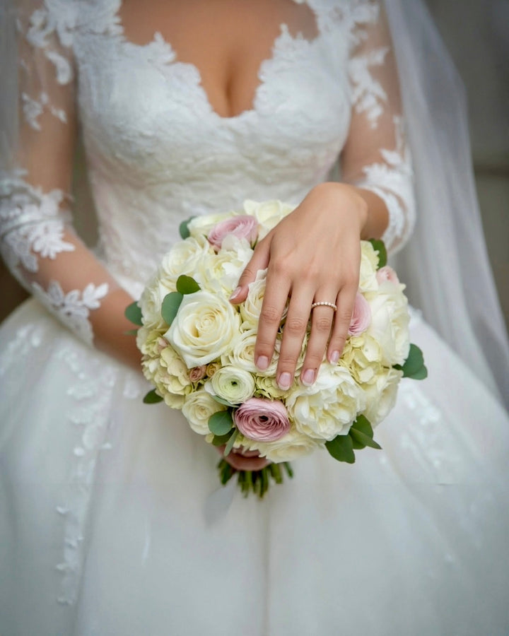Braut in elegantem Spitzenhochzeitskleid hält einen Blumenstrauß aus weißen und zartrosa Rosen, Nahaufnahme der Hand mit feinem Diamantring, romantische Hochzeitsästhetik.