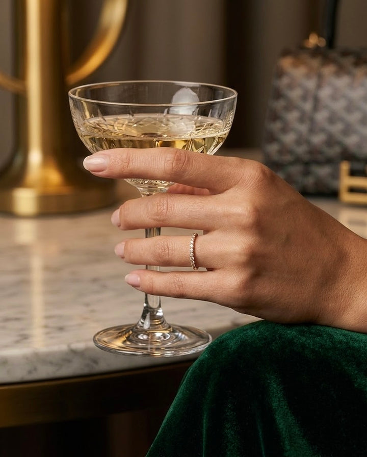 Feiner Damenring aus 18 Karat Gelbgold mit funkelnden weissen Zirkonia, getragen an einer Hand mit Champagnerglas in elegantem Ambiente.