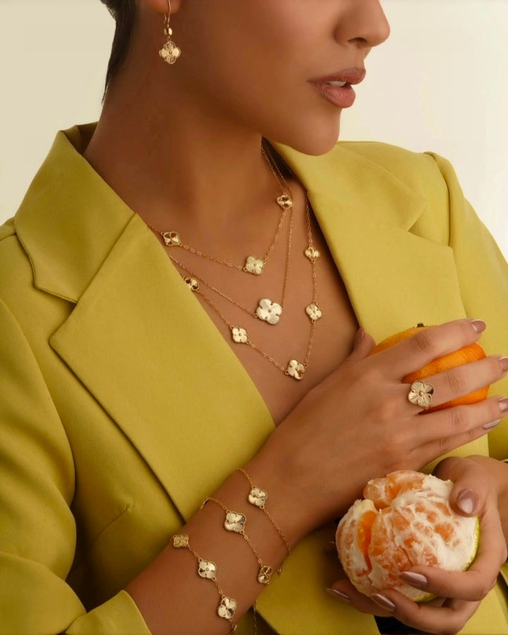 Detailaufnahme eines eleganten Models im grünen Blazer mit 18k-Gelbgoldschmuck: mehrreihige Halsketten, filigrane Ohrringe, Armband und Ring mit kleeblattförmigen Anhängern; stilvolle Inszenierung mit Zitrusfrüchten in der Hand.