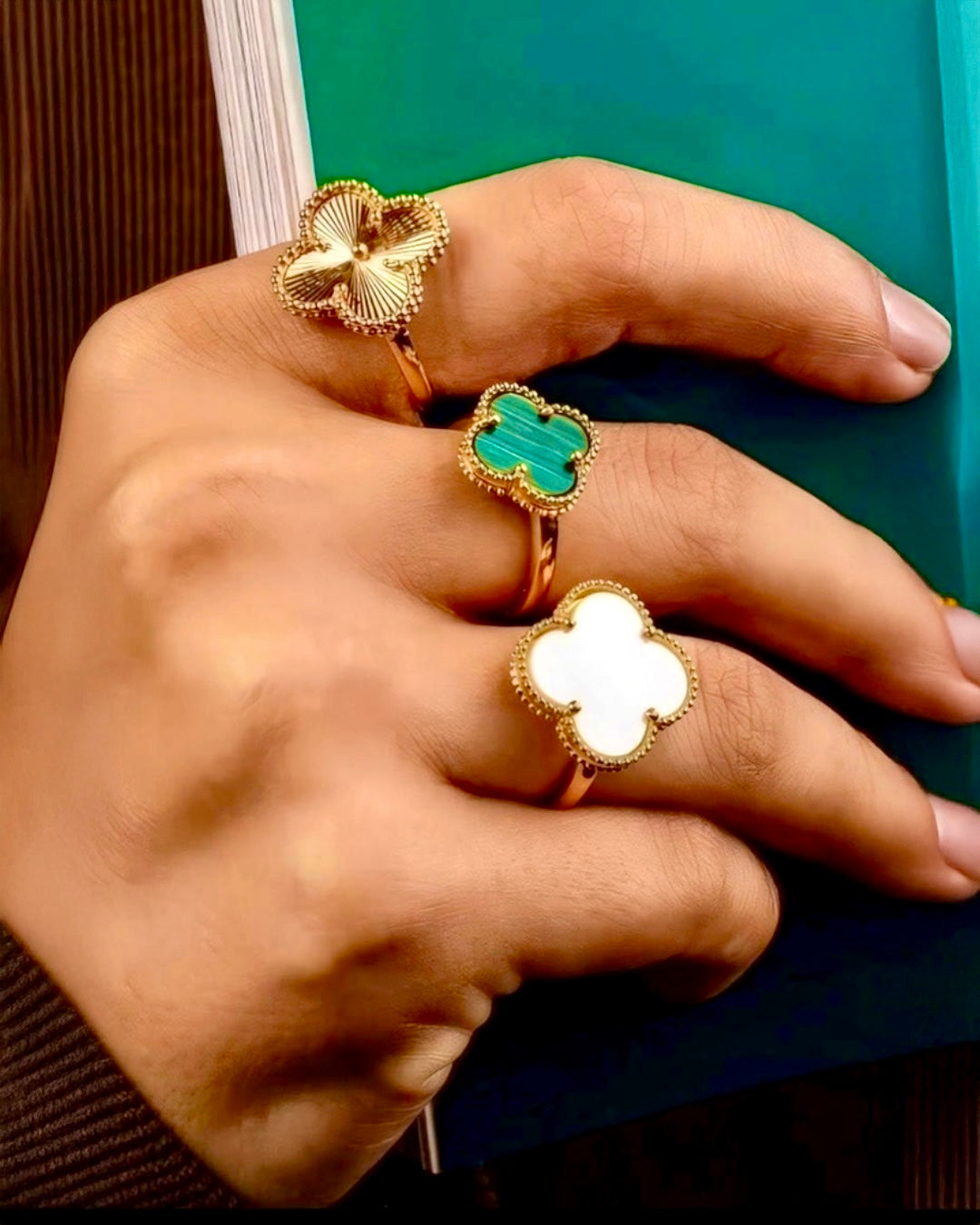Hand mit drei eleganten 18k-Gelbgoldringen im Kleeblatt-Design: ein Ring mit hochglanzpoliertem Goldmotiv, ein Ring mit grünem Edelstein-Einsatz und ein Ring mit weissem Perlmutt, kunstvoll gefasst, stilvoll arrangiert vor grünem Hintergrund.