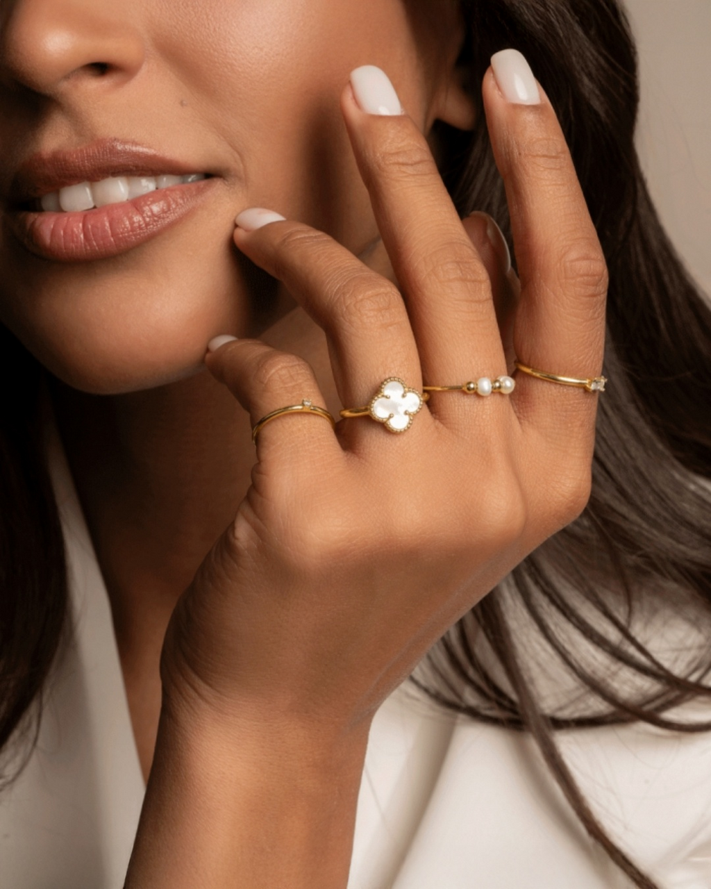 Nahaufnahme einer Frauenhand mit mehreren filigranen Ringen aus 18k Gelbgold: ein Ring mit kleinem Kleeblattmotiv aus weissem Perlmutt, ein zarter Perlenring sowie feine Goldringe, elegant getragen vor neutralem Hintergrund.