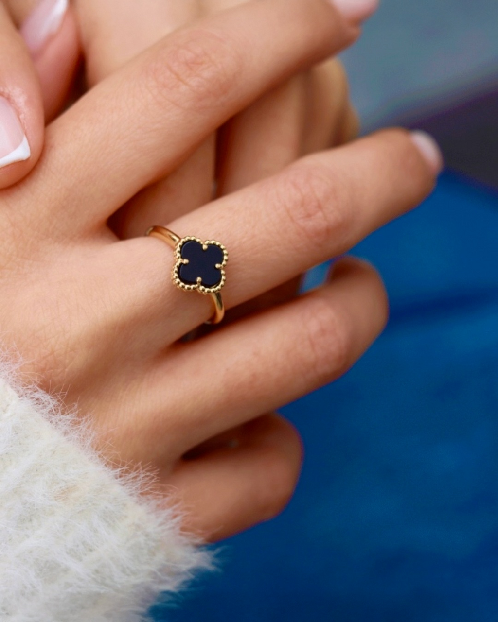 Ring aus 18k Gelbgold mit schwarzem Onyx in Kleeblattform, filigrane Goldfassung, getragen an der Hand, elegante Detailaufnahme vor blauem Hintergrund.