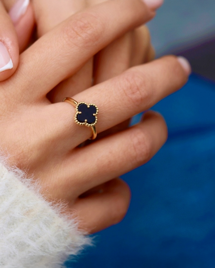 Ring aus 18k Gelbgold mit schwarzem Onyx in Kleeblattform, filigrane Goldfassung, getragen an der Hand, elegante Detailaufnahme vor blauem Hintergrund.