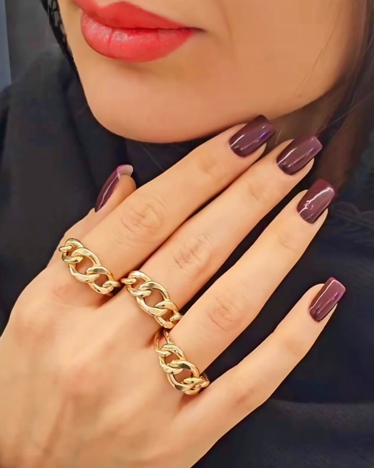 Nahaufnahme einer Frau mit rot geschminkten Lippen und dunkel lackierten Nägeln, die zwei auffällige Ringe aus 18 Karat Gelbgold mit markantem Kettenglieder-Design an der Hand trägt. Luxuriöser, moderner Statement-Look.