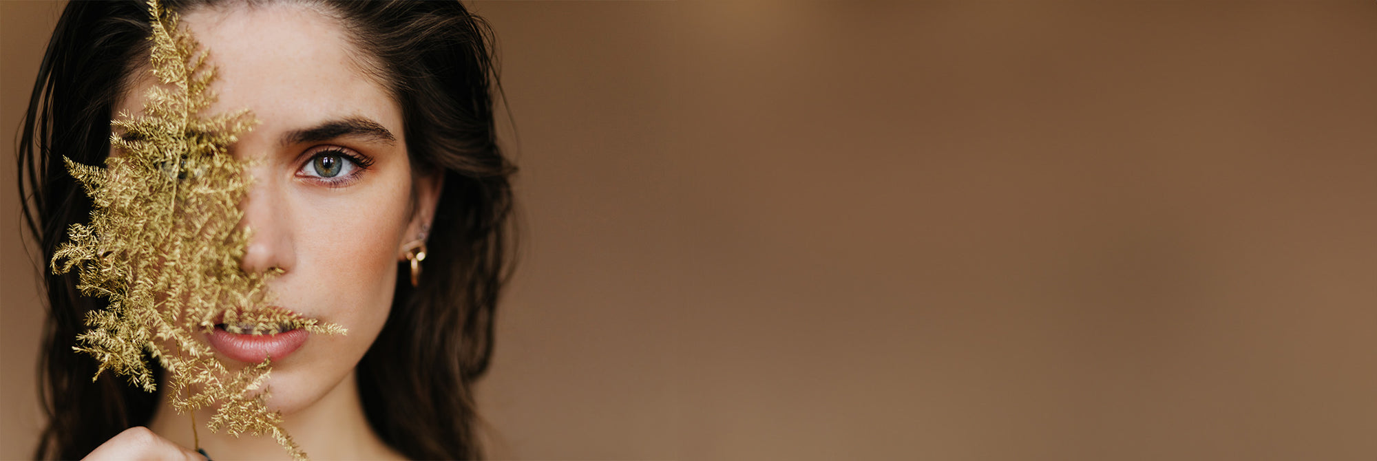 Hübsche junge Frau mit dunklen Haaren, goldener Ohrring, hält eine filigrane, goldüberzogene Pflanze vor die rechte Gesichtshälfte.